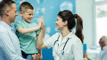 Niño feliz en la consulta de su doctora chocandole la mano mientras es sostenido por los brazos de su padre