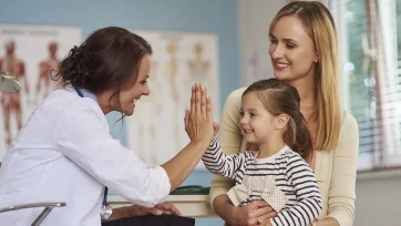 Niña contenta chocando las manos a su doctora en la consulta sujetada por los brazos de su madre feliz