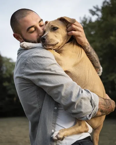 Joven abrazando en brazos a un perro de gran tamaño en una explanada con arboles al fondo