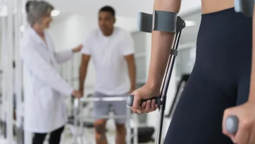 Joven con muletas en primer plano en una sala de rehabilitacion y al fondo difuminados un doctor con el paciente hablando