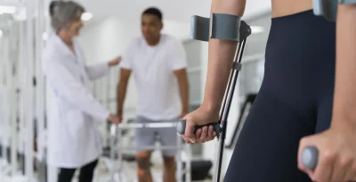 Joven con muletas en primer plano en una sala de rehabilitacion y al fondo difuminados un doctor con el paciente hablando